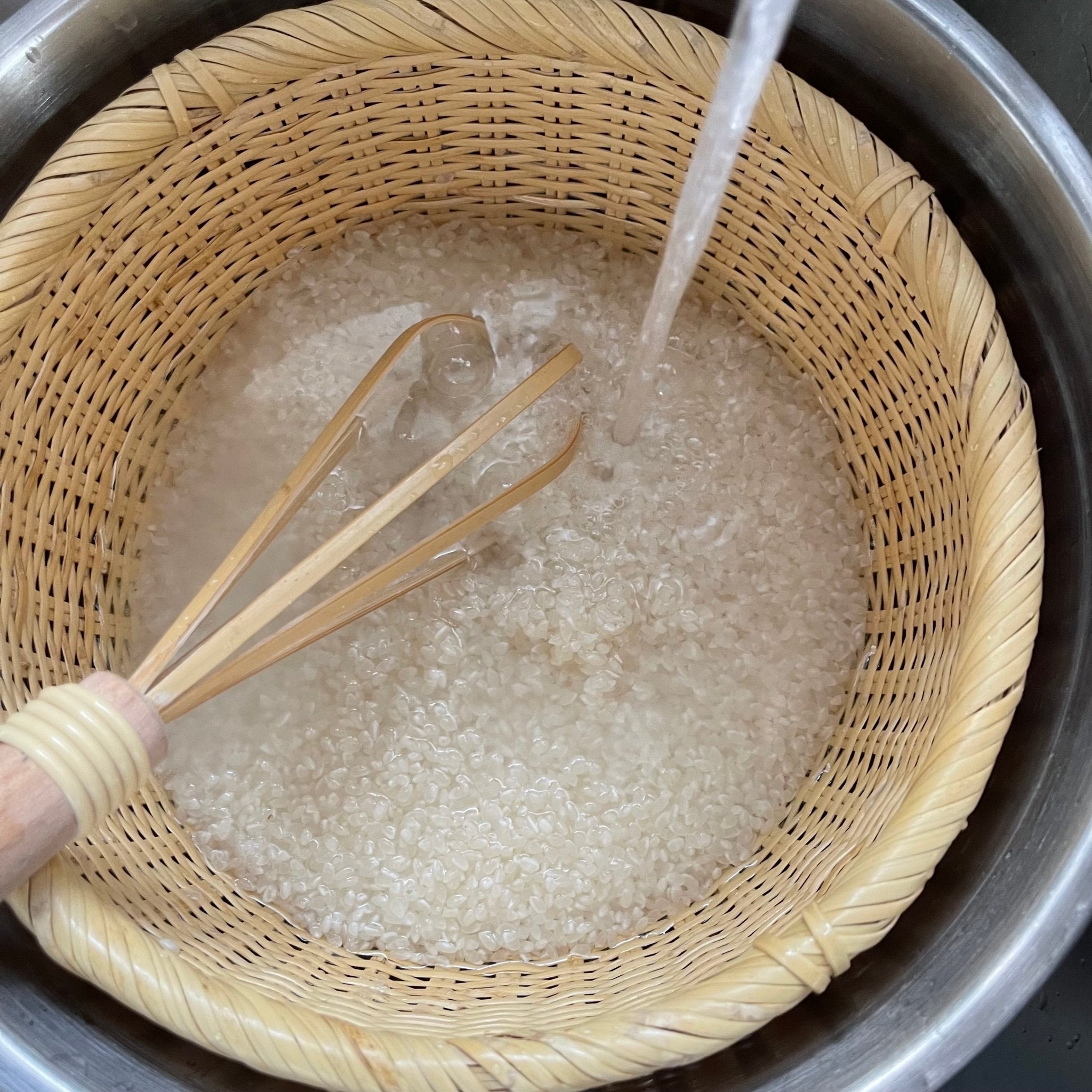 Close-up of bamboo rice washing whisk stirring white rice in a woven strainer basket under running water, demonstrating traditional Japanese rinsing technique.