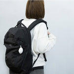 Woman carrying a black backpack with the Cocoon Eco Bag by 100 Percent attached to the front zipper. The compact, oval-shaped white case hangs from the bag, highlighting its convenient clip-on design and stylish minimalism for everyday travel or errands.