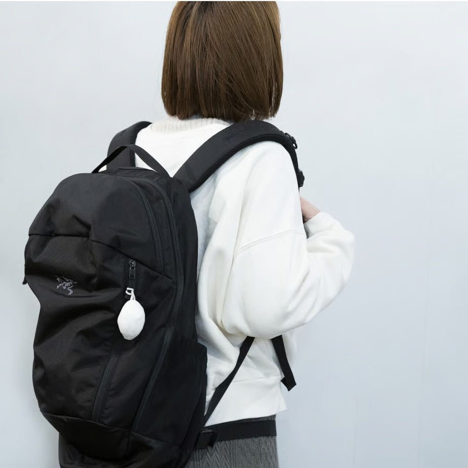 Woman carrying a black backpack with the Cocoon Eco Bag by 100 Percent attached to the front zipper. The compact, oval-shaped white case hangs from the bag, highlighting its convenient clip-on design and stylish minimalism for everyday travel or errands.
