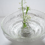Delicate white flowers held by a sculptural silver spiral flower holder submerged in a rippled glass bowl, showcasing a modern minimalist approach to ikebana and artistic floral design.