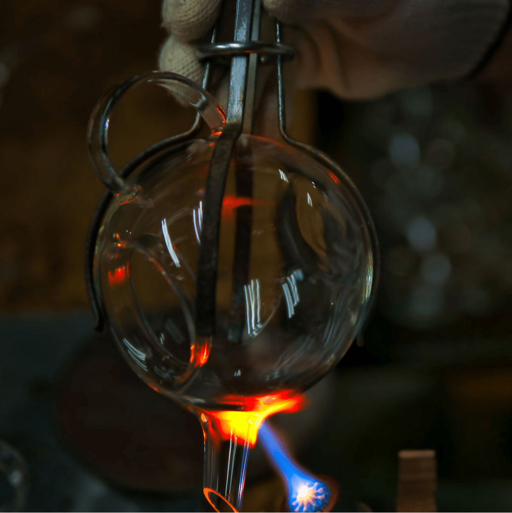 Artisan crafting a Manmaru glass teapot using traditional glassblowing techniques, showing the glowing molten glass being shaped by hand over an open flame.