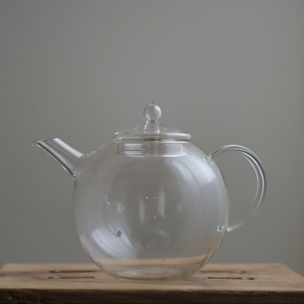 Transparent Manmaru glass teapot with a classic handle and round shape, placed on a wooden surface, ideal for serving tea while showcasing the natural colors of the brew.