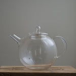 Transparent Manmaru glass teapot with a classic handle and round shape, placed on a wooden surface, ideal for serving tea while showcasing the natural colors of the brew.