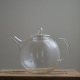 Transparent Manmaru glass teapot with a classic handle and round shape, placed on a wooden surface, ideal for serving tea while showcasing the natural colors of the brew.