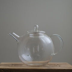 Transparent Manmaru glass teapot with a classic handle and round shape, placed on a wooden surface, ideal for serving tea while showcasing the natural colors of the brew.