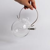 A clear view of the Manmaru glass teapot being held by hand, highlighting its handcrafted design, heat-resistant glass body, brass handle, and stainless steel filter—an elegant blend of Japanese craftsmanship and functionality.