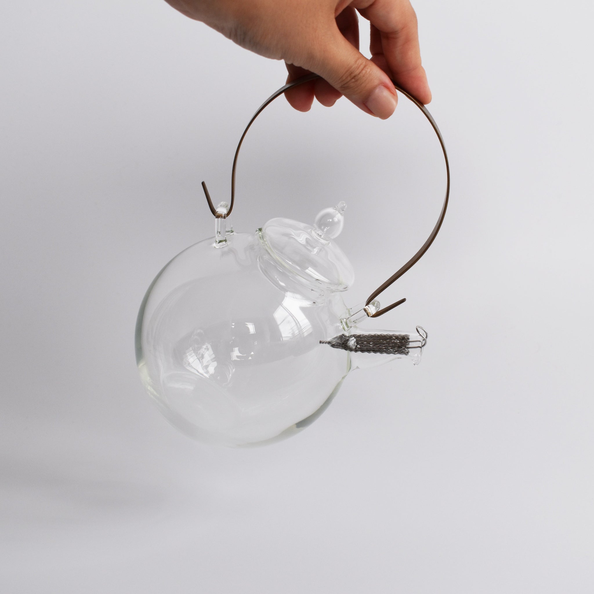 A clear view of the Manmaru glass teapot being held by hand, highlighting its handcrafted design, heat-resistant glass body, brass handle, and stainless steel filter—an elegant blend of Japanese craftsmanship and functionality.