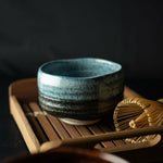Seifu blue hand-glazed matcha bowl placed on a wooden tray beside a bamboo whisk, evoking a traditional Japanese tea ceremony setting with rich texture and moody lighting.